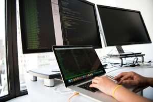 Home A developer writes code on a laptop in front of multiple monitors in an office setting.